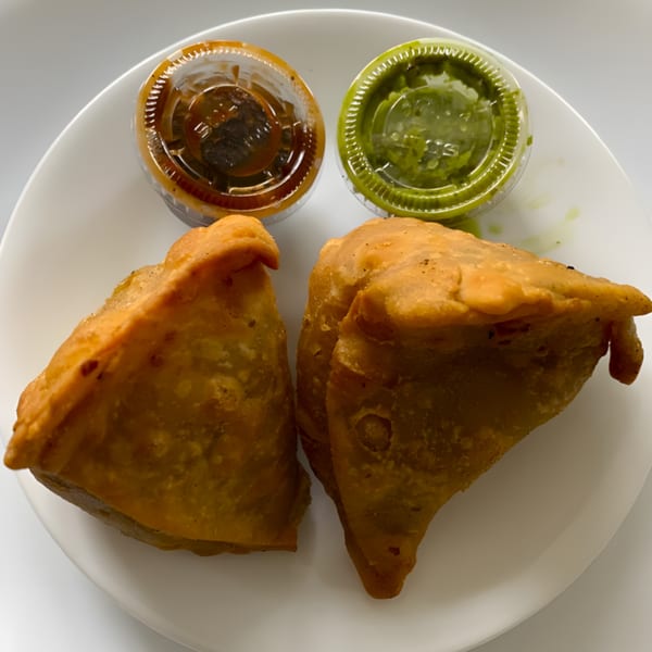 Two golden-brown Clay Oven samosas with small containers of green and brown chutneys on a white plate.